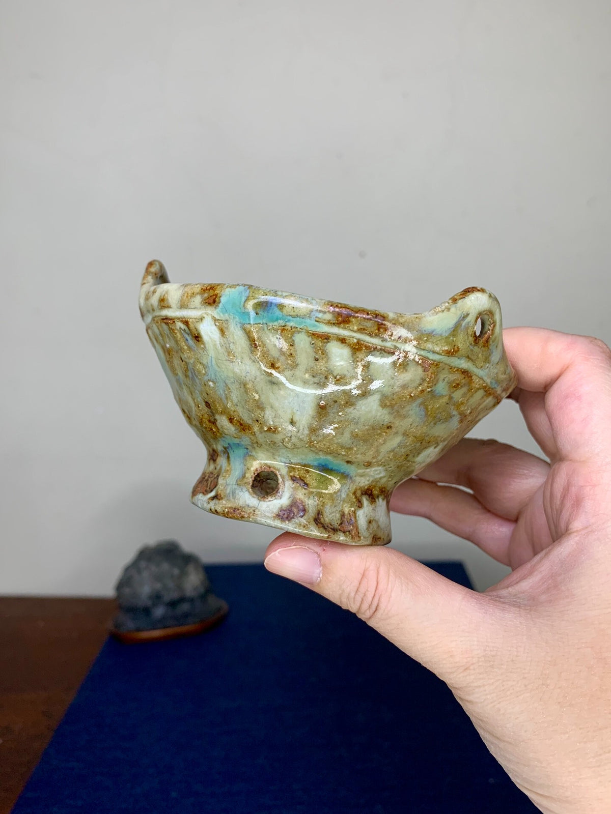 A hand holds a small, glazed ceramic pot with brown and blue glaze drips and a textured rim.