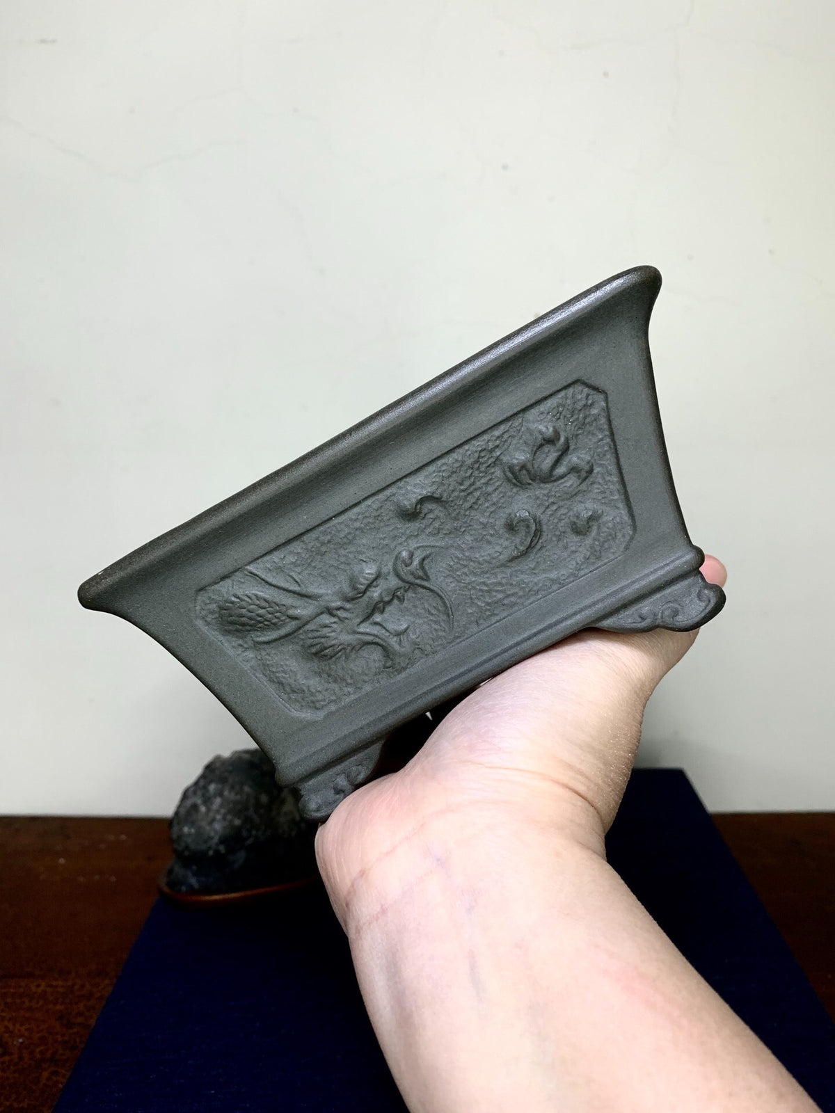 A hand holds a dark grey, rectangular pot with an embossed floral design on its side.