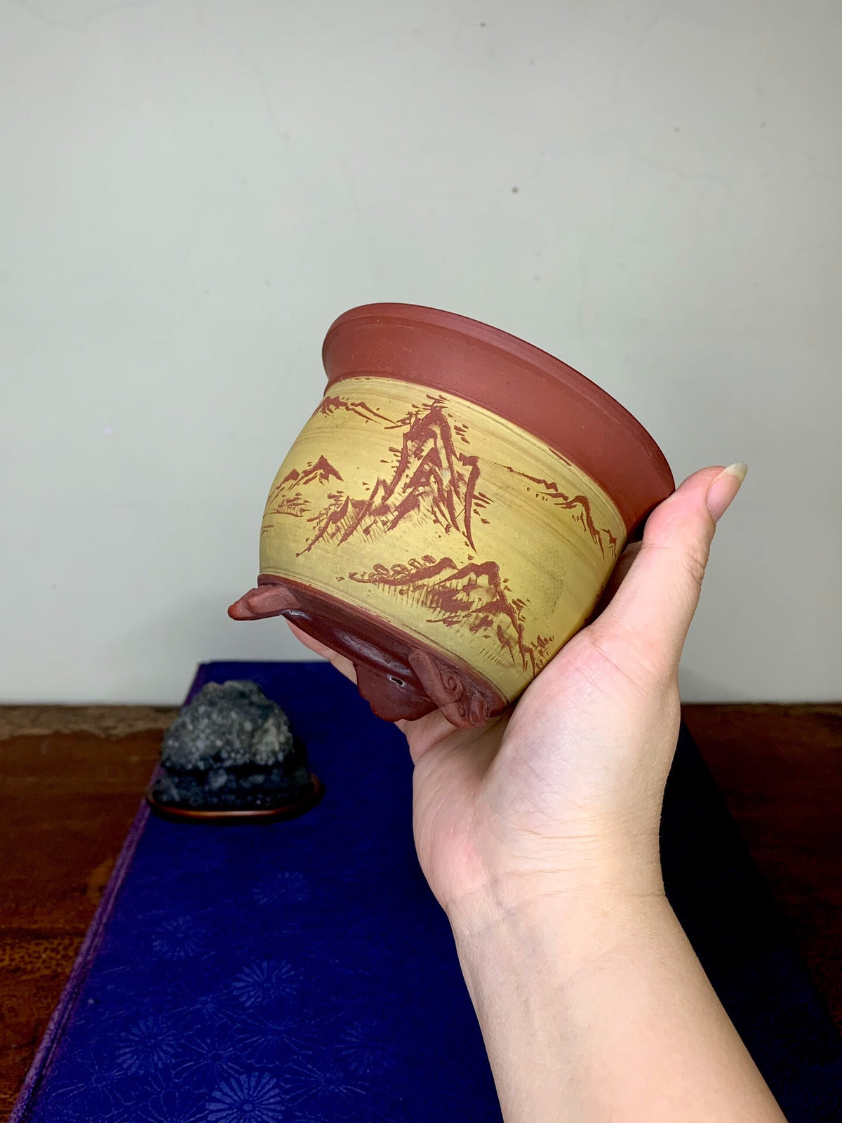 A hand holds a small, reddish-brown ceramic pot with a yellow glazed band featuring red mountain scenery.