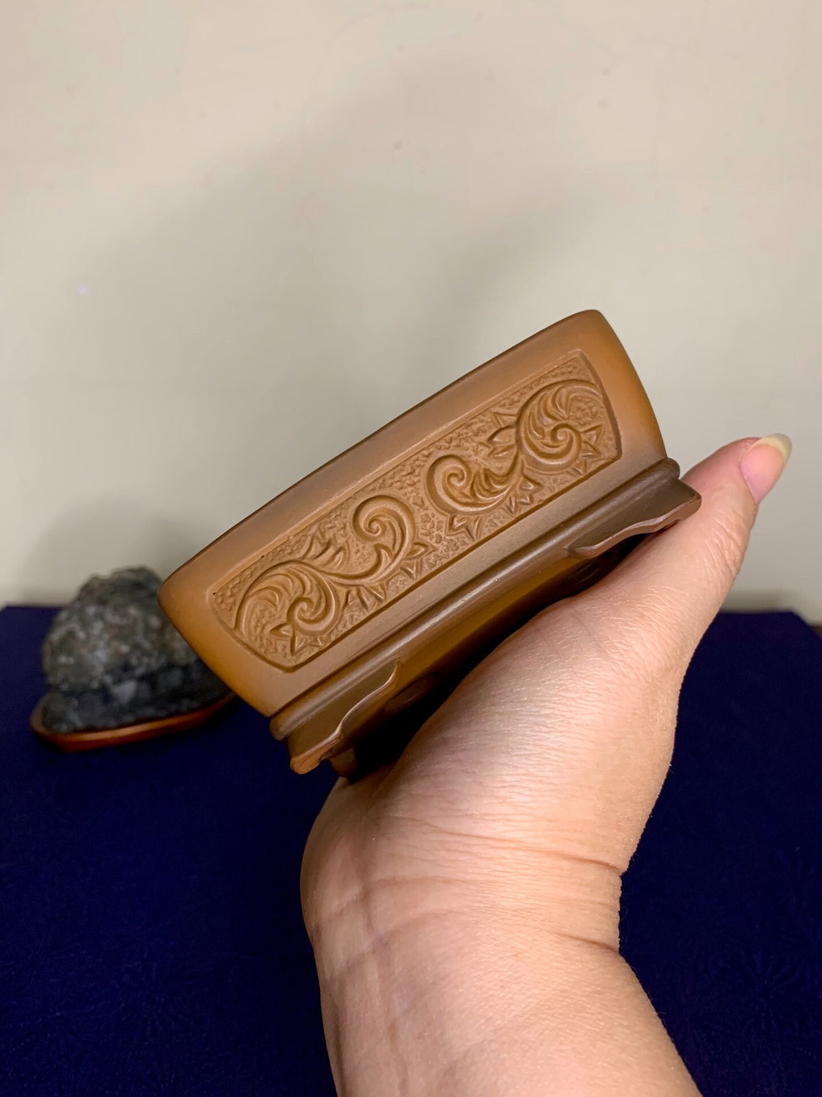 A hand holds a brown glazed rectangular pot with carved swirling patterns on its side.