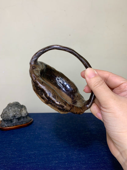 A hand holds a dark brown, glossy, irregularly shaped ceramic pot with a looped handle.