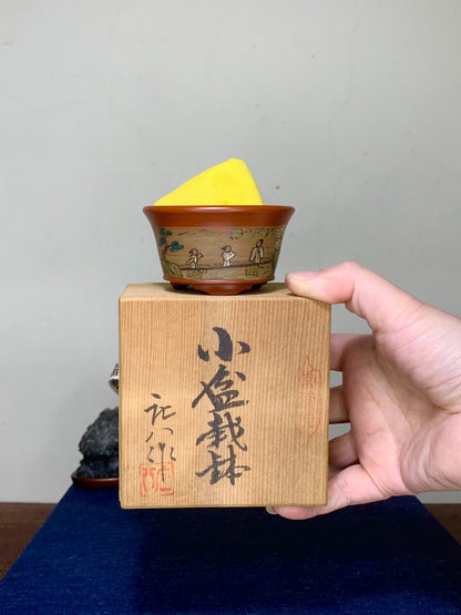 Hand holds wooden box with black calligraphy, topped by small brown glazed pot with yellow lid and painted figures.