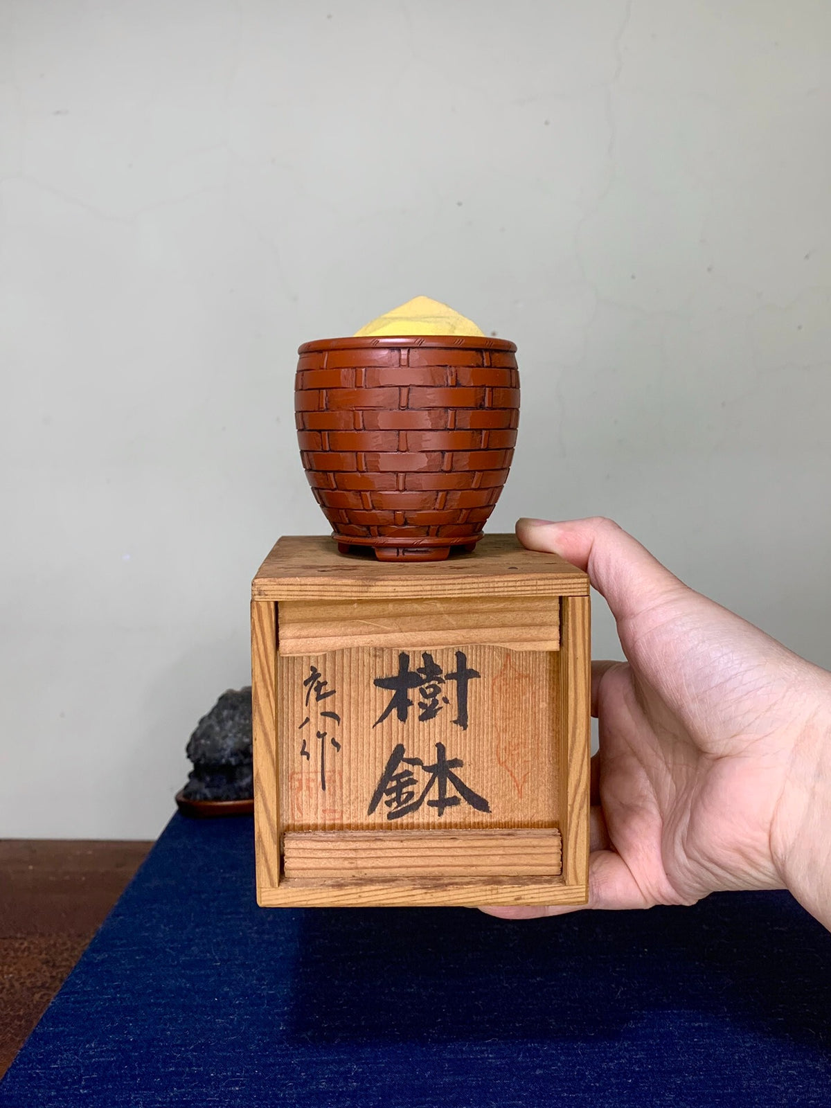 A hand holds a wooden box with black Japanese characters, topped by a brown, woven-textured pot with a yellow lid.