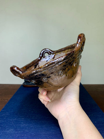 A hand holds a glossy brown glazed ceramic pot with two loop handles and three circular marks.