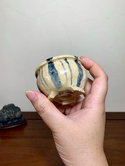 A hand holds a small, beige ceramic pot with blue brushstroke patterns and three feet.