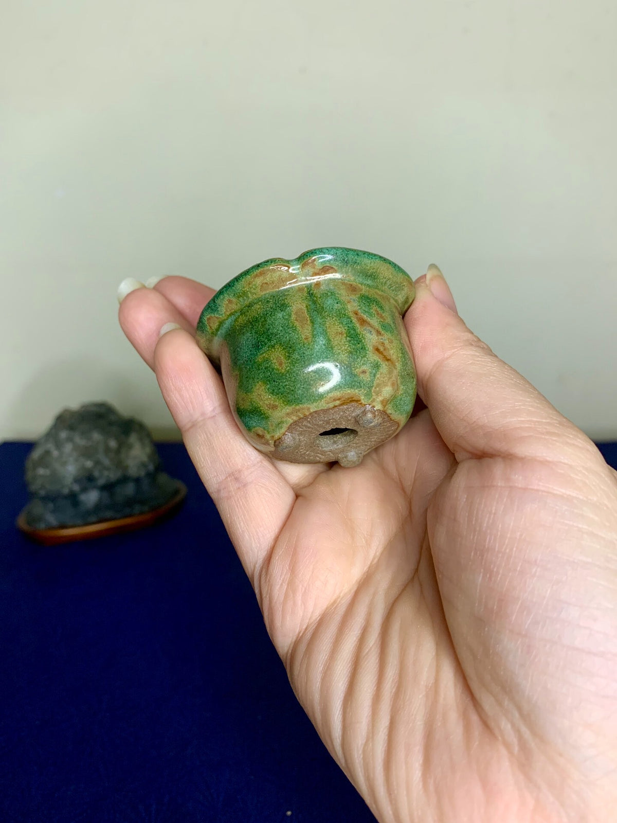 A hand holds a small, rounded green glazed pot with brown mottling and a central drainage hole.