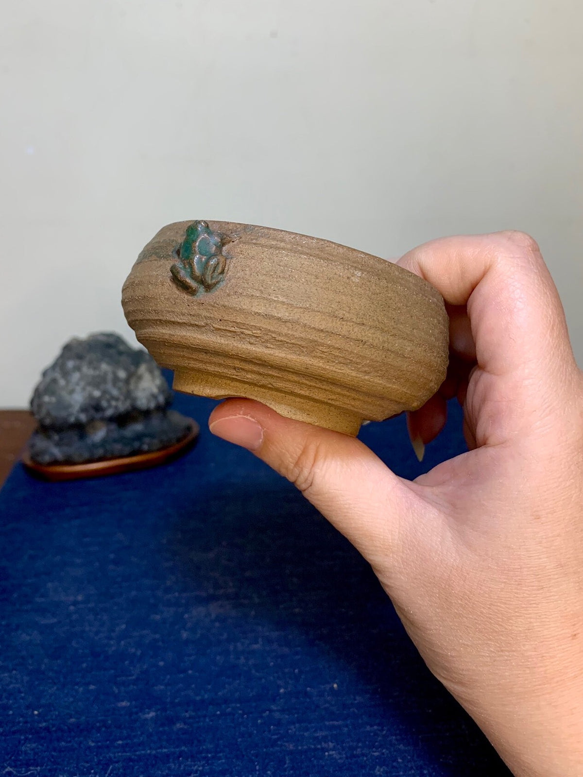 A hand holds a small, brown, unglazed ceramic pot with a green frog decoration on its rim.