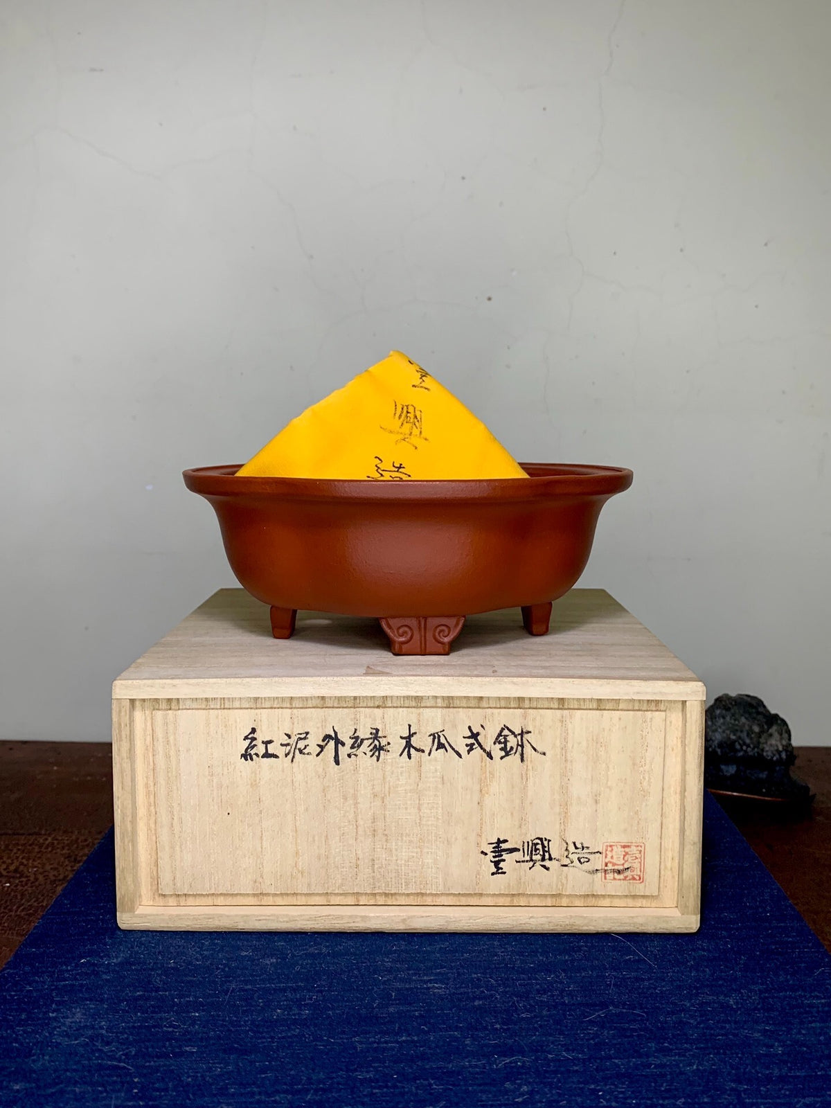 A small, reddish-brown, unglazed ceramic pot with three feet, covered by a yellow paper cone.