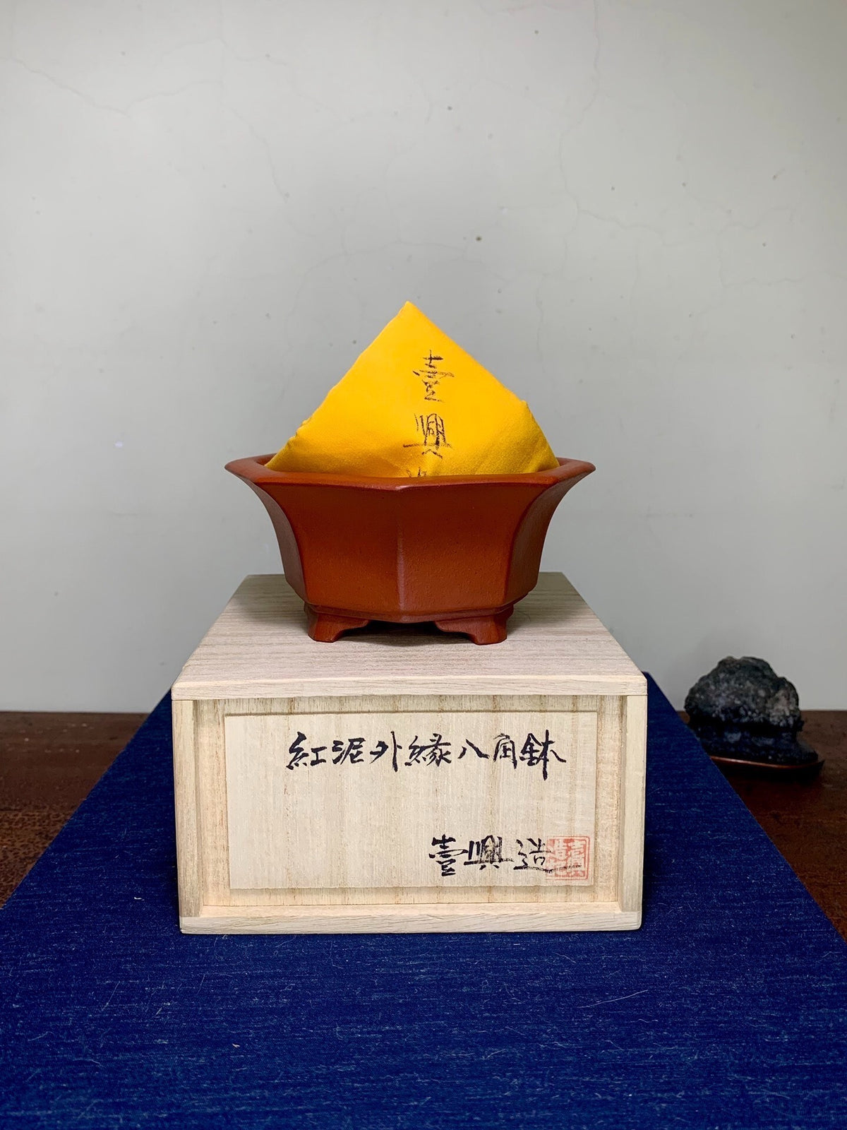 A small, reddish-brown, octagonal ceramic pot with a yellow paper label sits atop a wooden box.