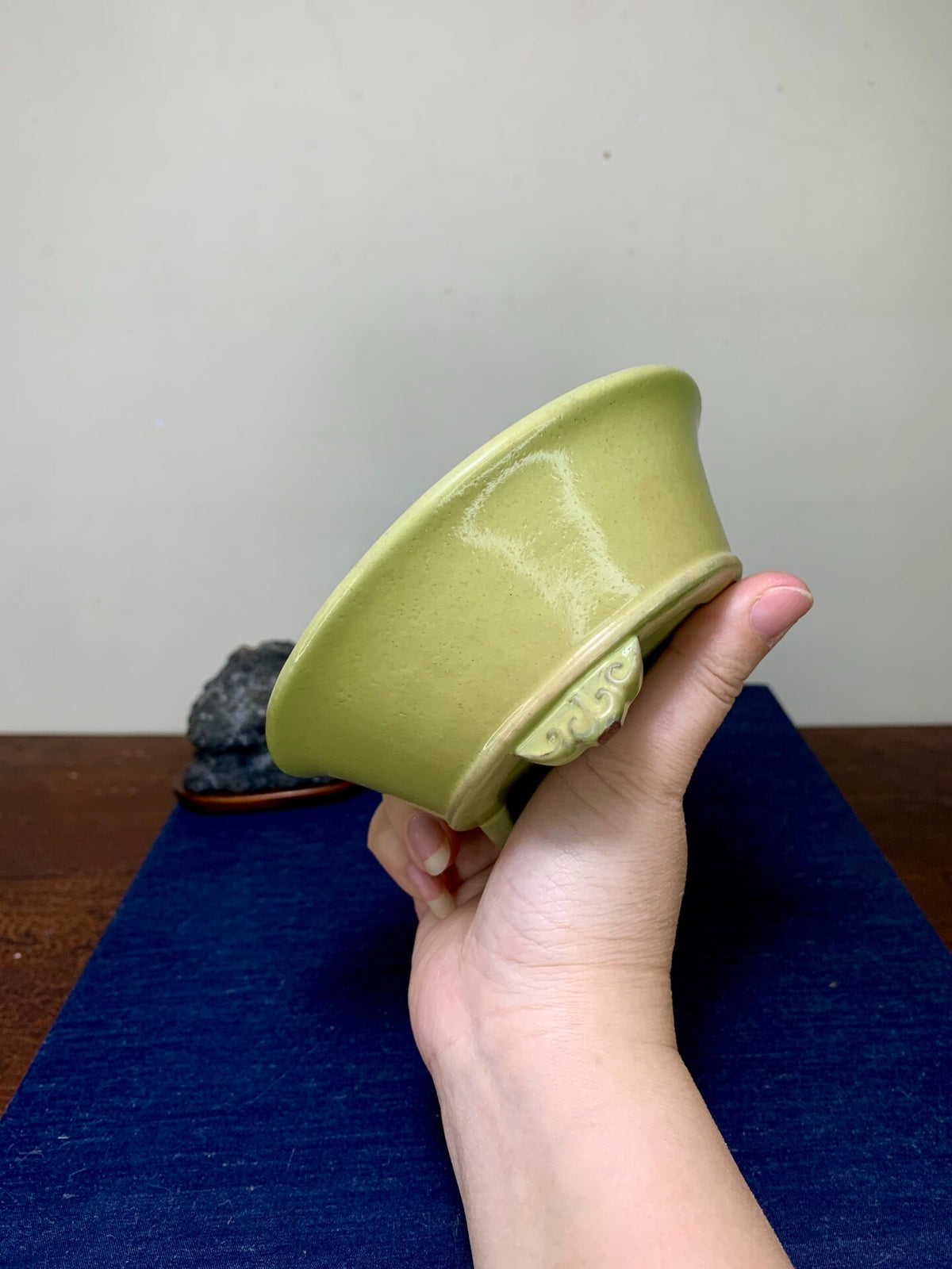 A hand holds a small, light green glazed ceramic pot with a flared rim and embossed markings.