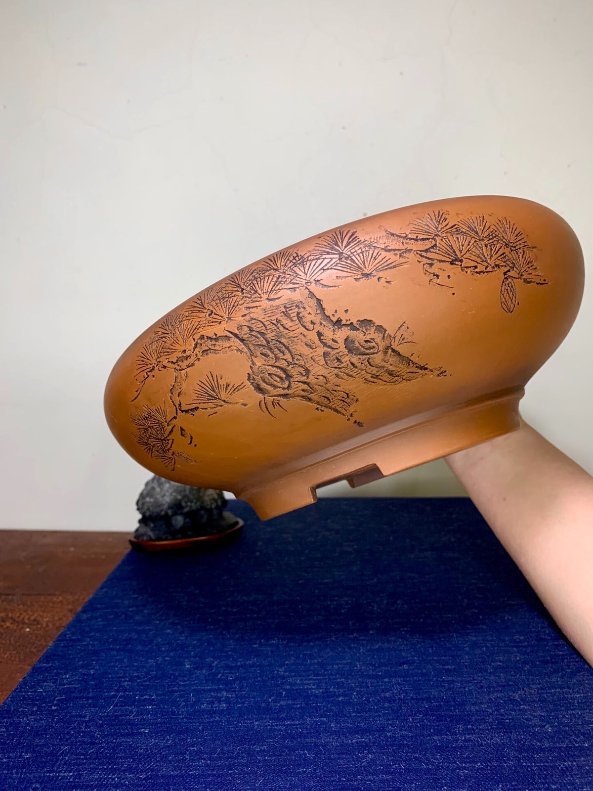 A hand holds a brown glazed pot with engraved pine and rock design.