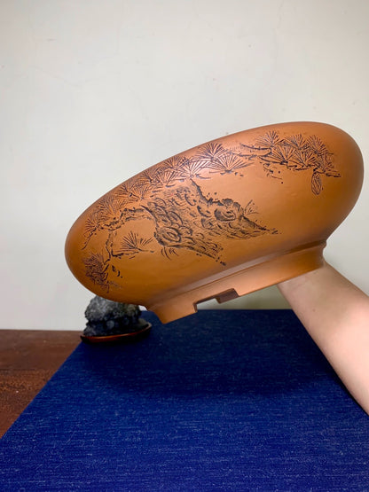 A hand holds a brown glazed pot with engraved pine and rock design.