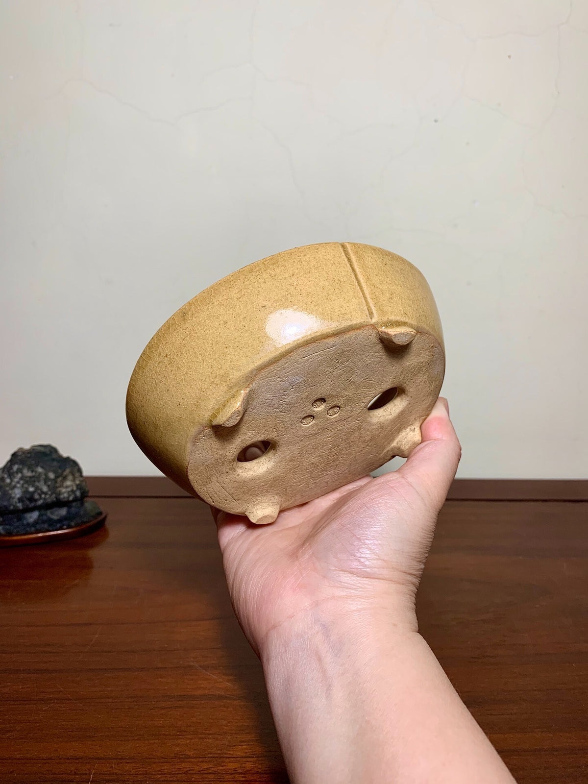 A hand holds a small, beige, unglazed ceramic pot with four feet and drainage holes.