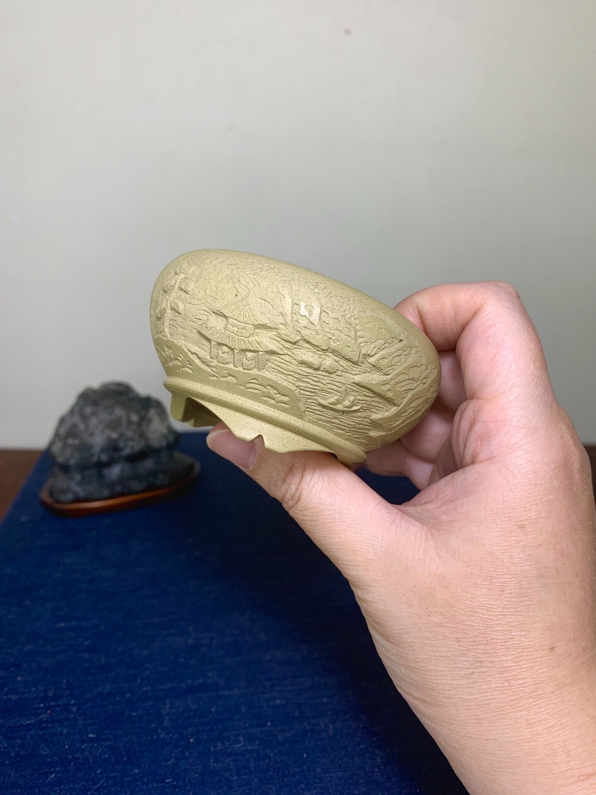 A hand holds a pale yellow, matte, intricately carved bonsai pot with relief mountain and tree designs.