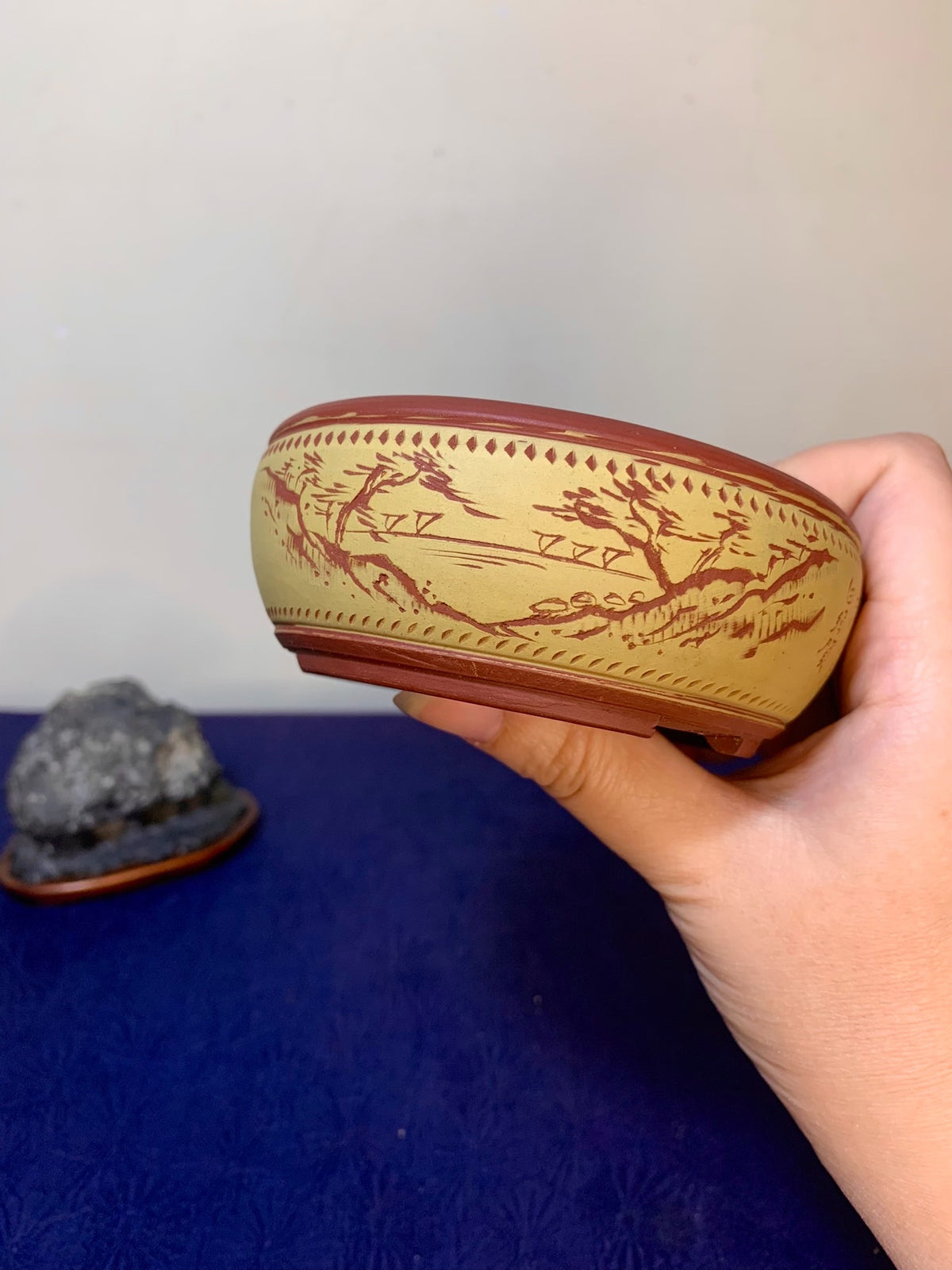 A hand holds a beige ceramic pot with reddish-brown painted landscape and decorative border.