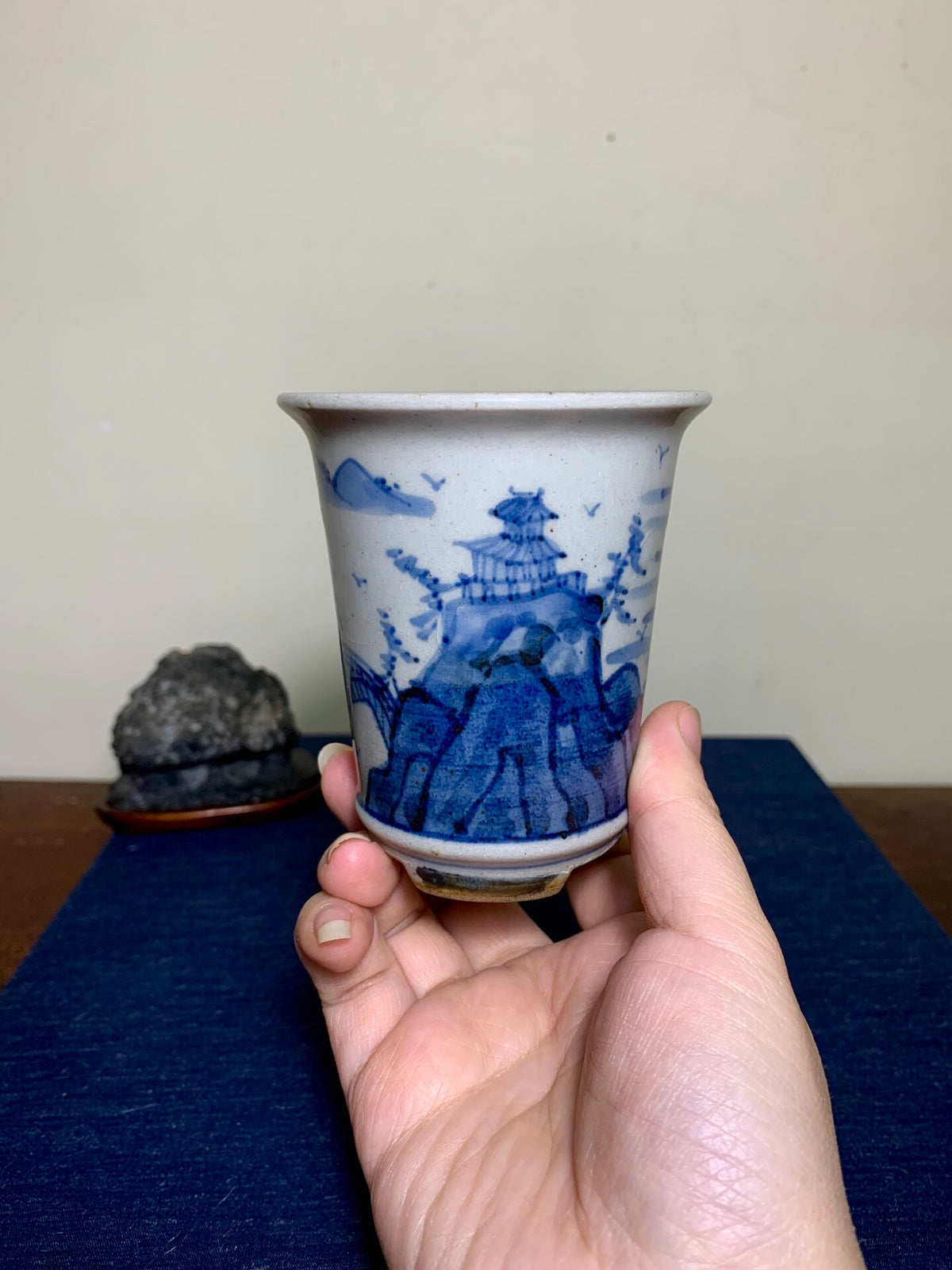 A hand holds a small, white glazed pot with blue painted landscape and pagoda design.