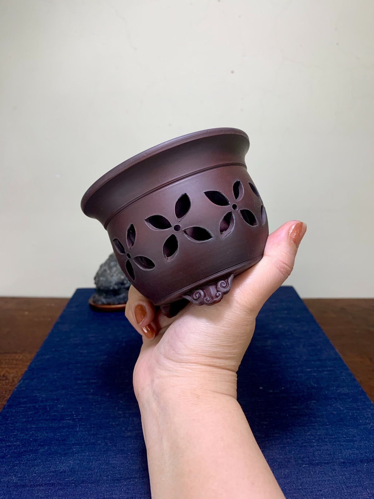 A hand holds a dark brown ceramic pot with floral cutouts and a small decorative foot.