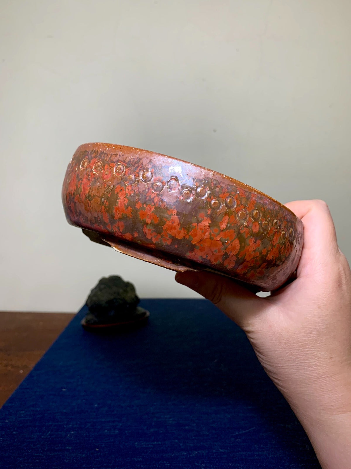 A hand holds a round, glossy reddish-brown pot with orange speckles and a textured rim.