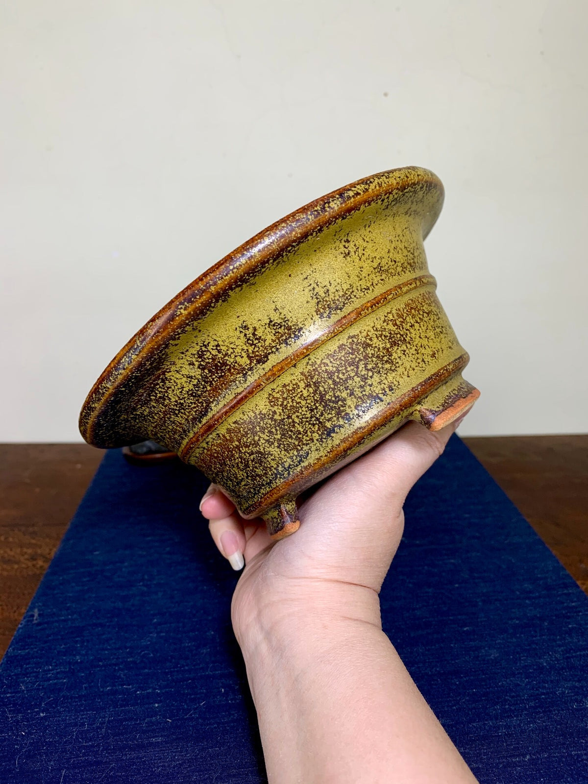 A hand holds a glazed ceramic pot with a yellowish glaze and brown speckles and rim.