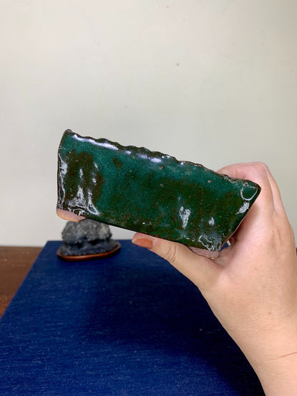 A hand holds a glossy, dark green rectangular ceramic pot with a rough, uneven rim.