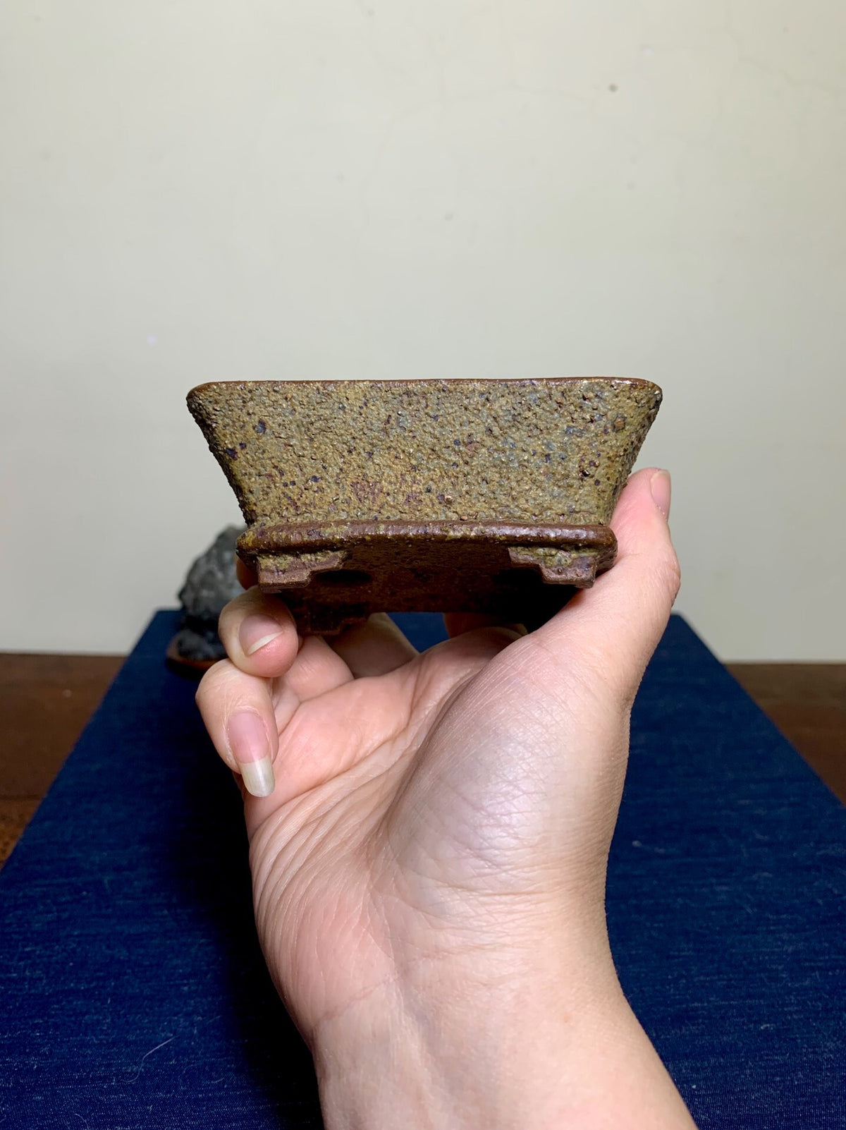A hand holds a small, rectangular, unglazed ceramic pot with a speckled brown and grey surface.