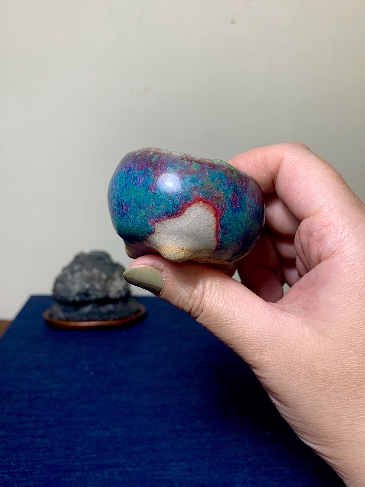 A hand holds a small, rounded pot with a glossy, multicolored glaze in blue, red, and white.