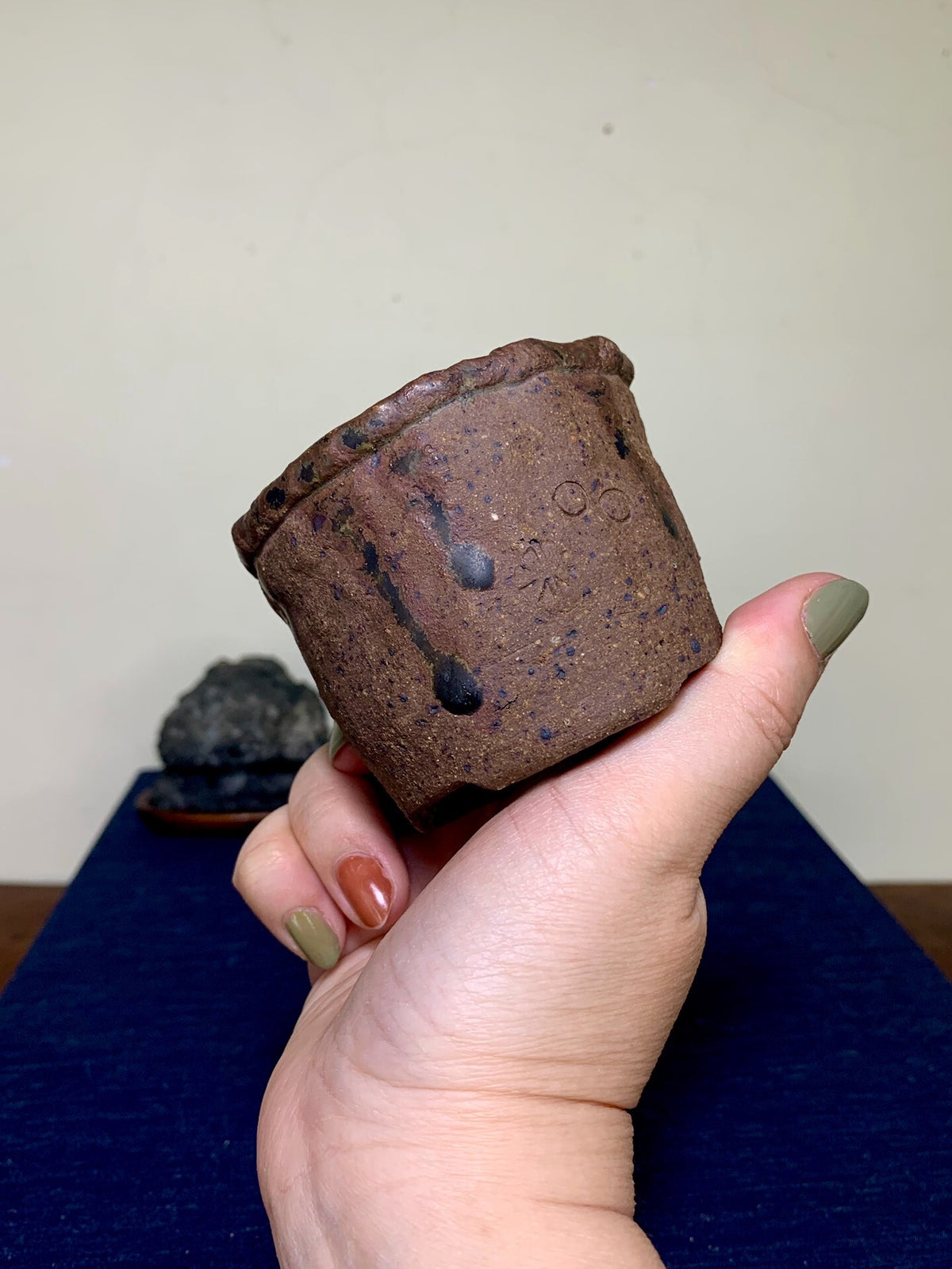 A hand holds a small, dark brown, unglazed ceramic pot with black speckles and a rough texture.
