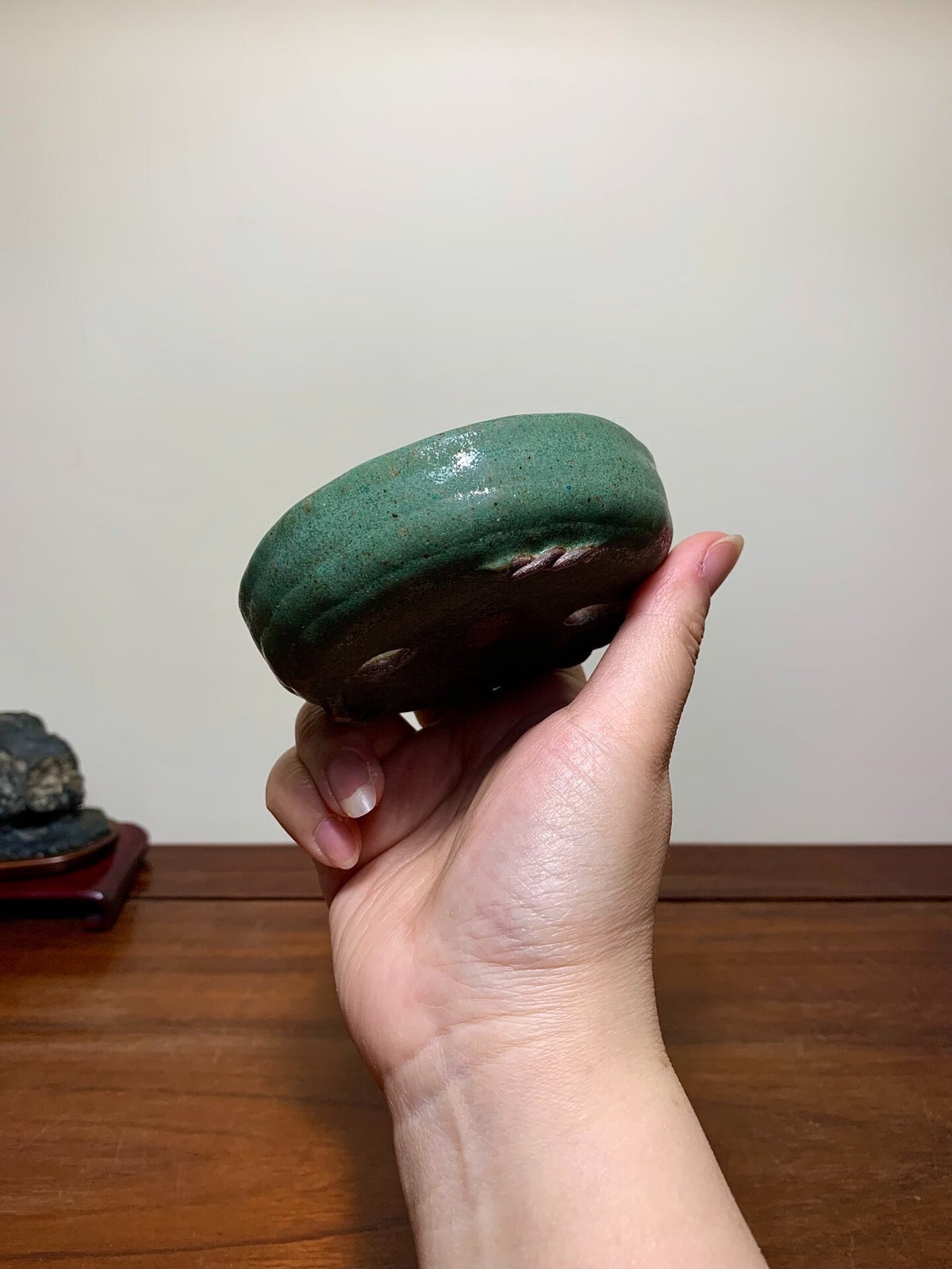 A hand holds a small, round, glossy green glazed pot with a speckled surface.