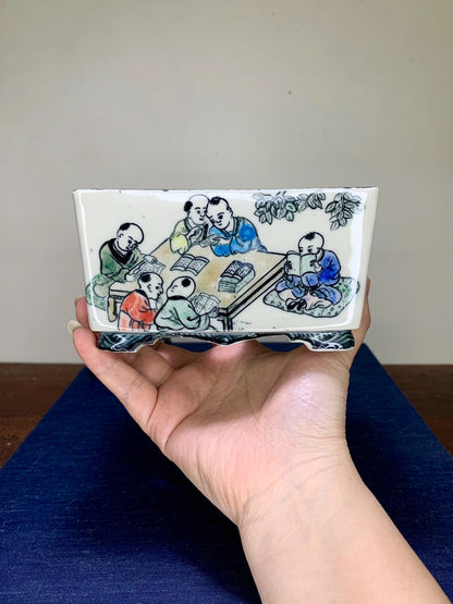 A hand holds a rectangular glazed ceramic pot with a painted scene of children studying.