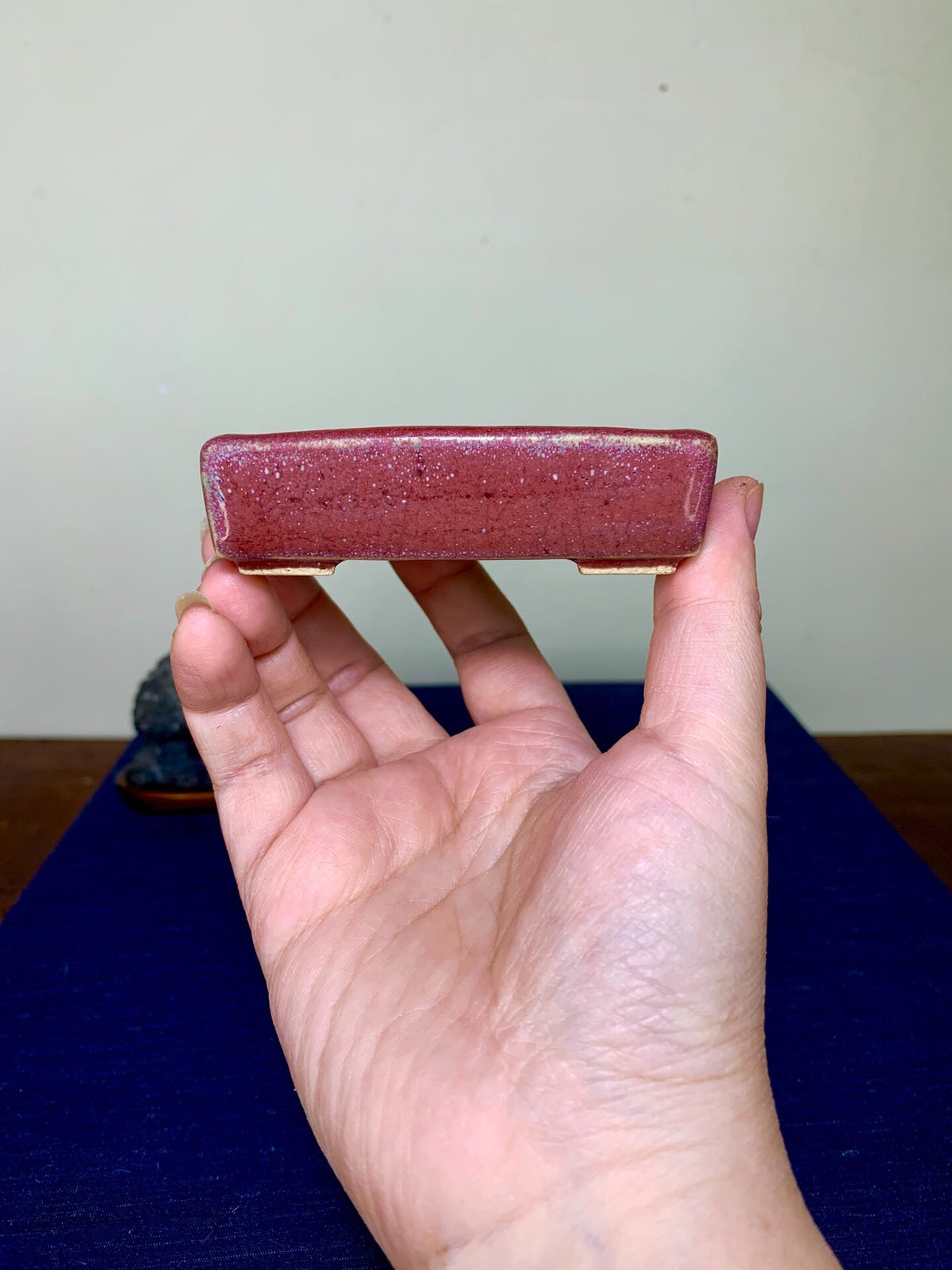 A hand holds a small, rectangular, deep red glazed pot with a speckled surface.