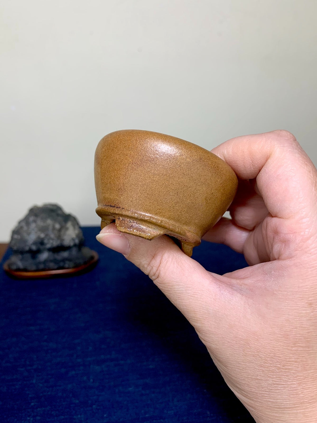 A hand holds a small, glossy brown ceramic pot with a rounded body and three short feet.