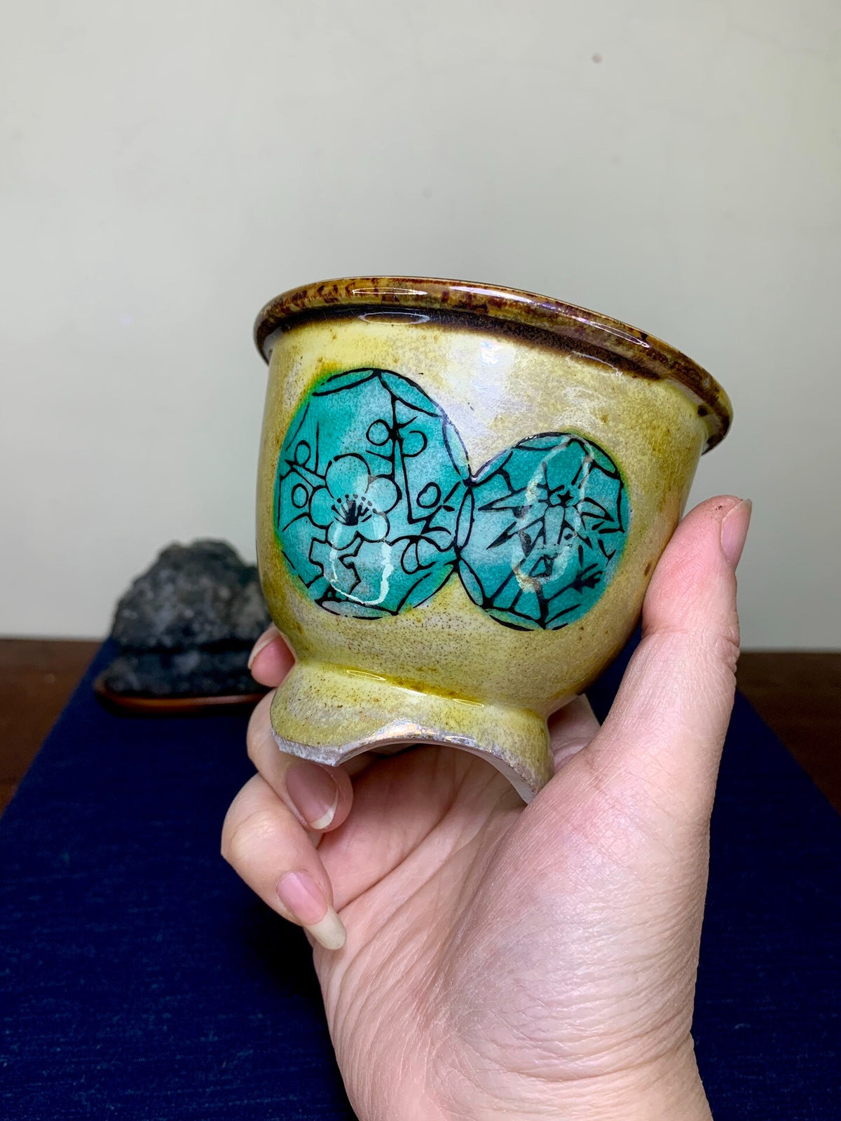 A hand holds a small, yellow glazed pot with two turquoise floral designs outlined in black.