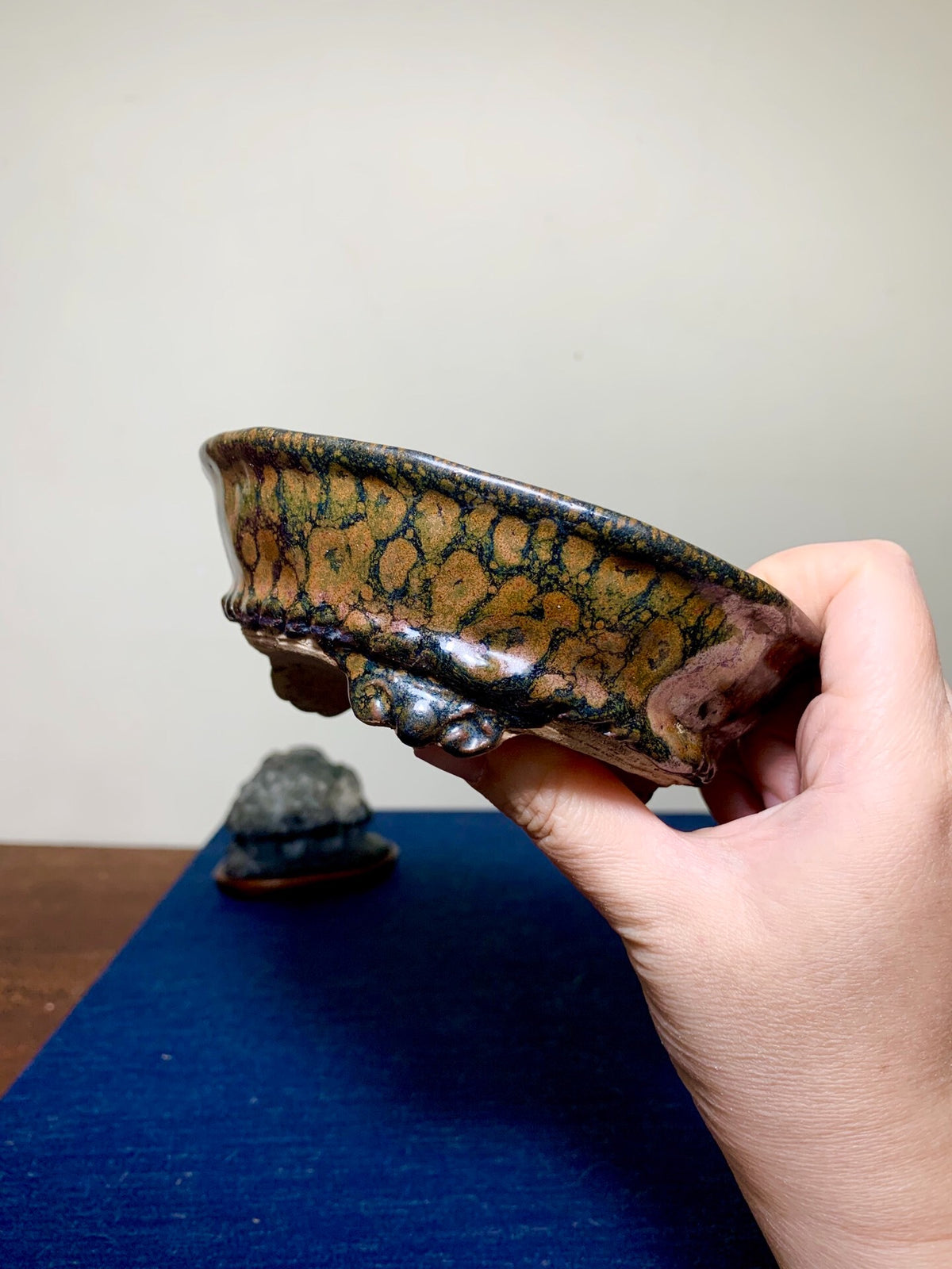 Hand holds a glazed ceramic pot with mottled brown and green glaze, shallow bowl shape.
