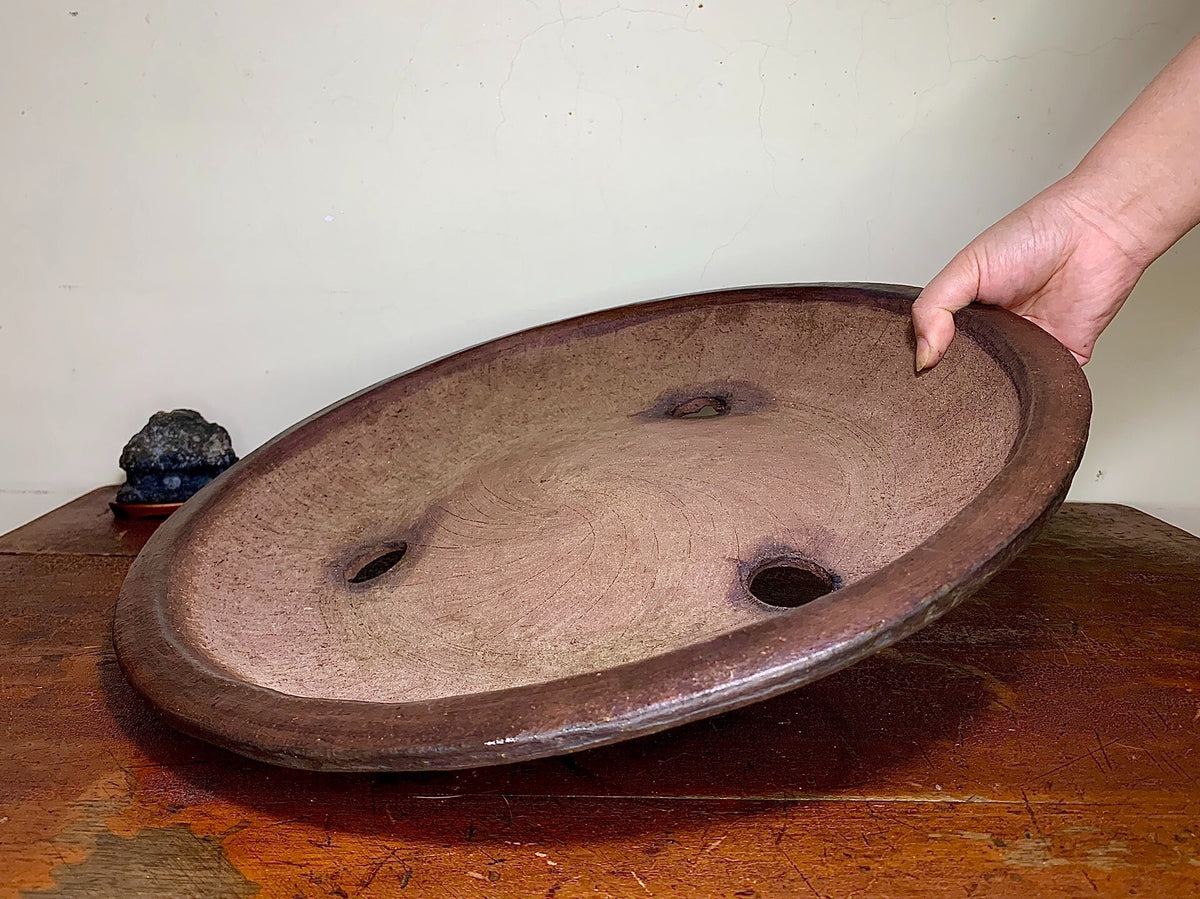 A hand holds a large, shallow, oval wooden tray with three holes and a worn, dark brown surface.