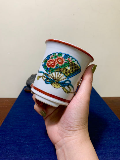 A hand holds a white ceramic cup with a red rim, featuring a colorful fan and floral design.