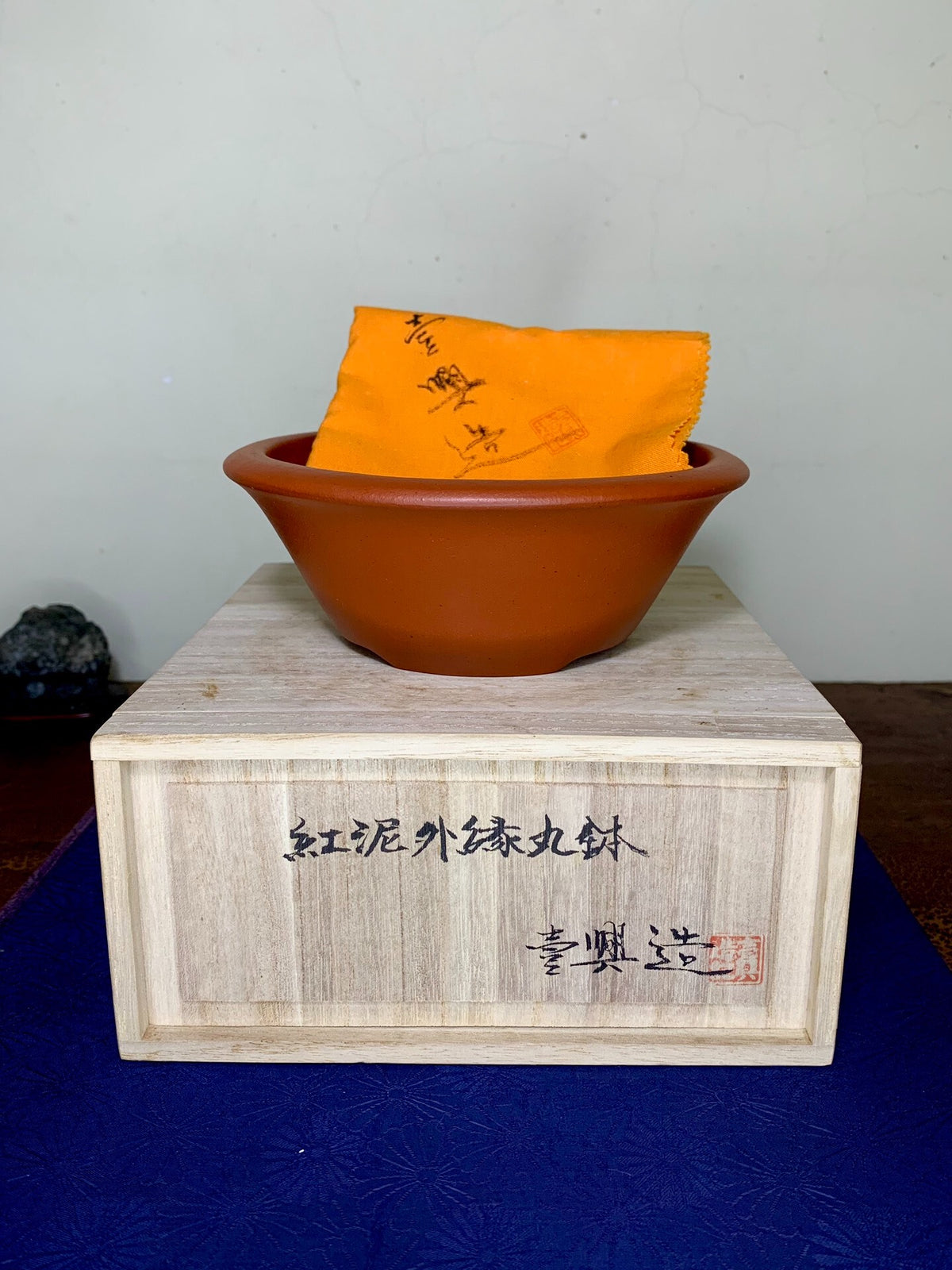A terracotta bonsai pot with a wide rim, resting on a wooden box with Japanese characters.