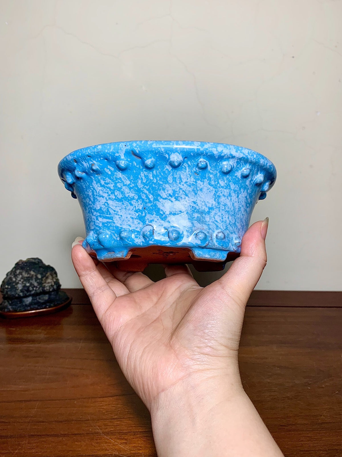 A hand holds a blue glazed pot with raised bumps and a brown base.