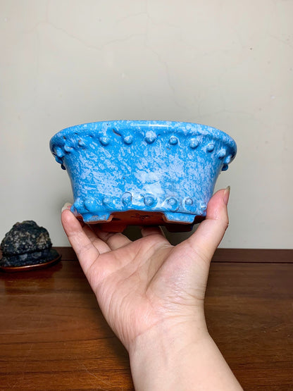 A hand holds a blue glazed pot with raised bumps and a brown base.