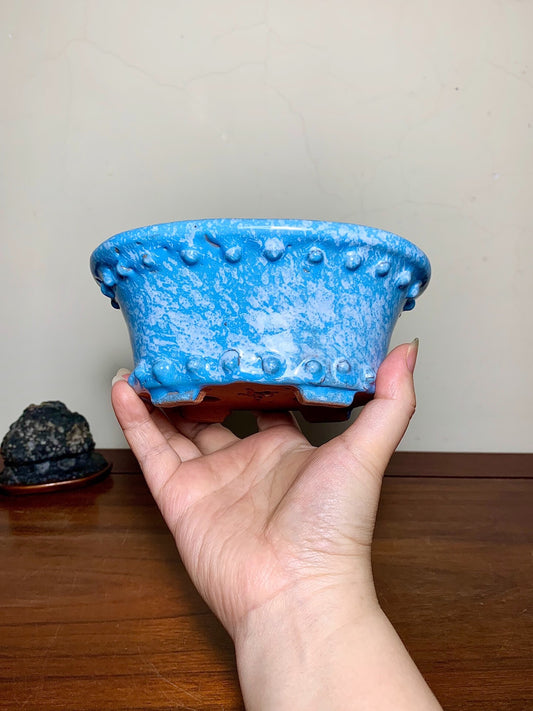 A hand holds a blue glazed pot with raised bumps and a brown base.