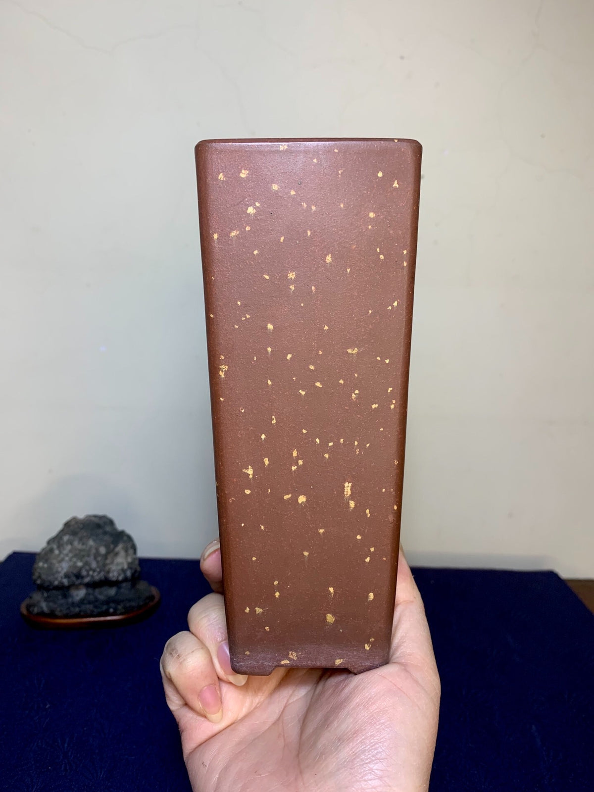 A hand holds a tall, rectangular brown pot with scattered gold flecks on a matte surface.
