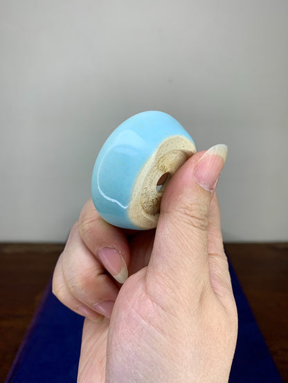 A hand holds a small, light blue glazed pot with a beige unglazed rim and a central hole.