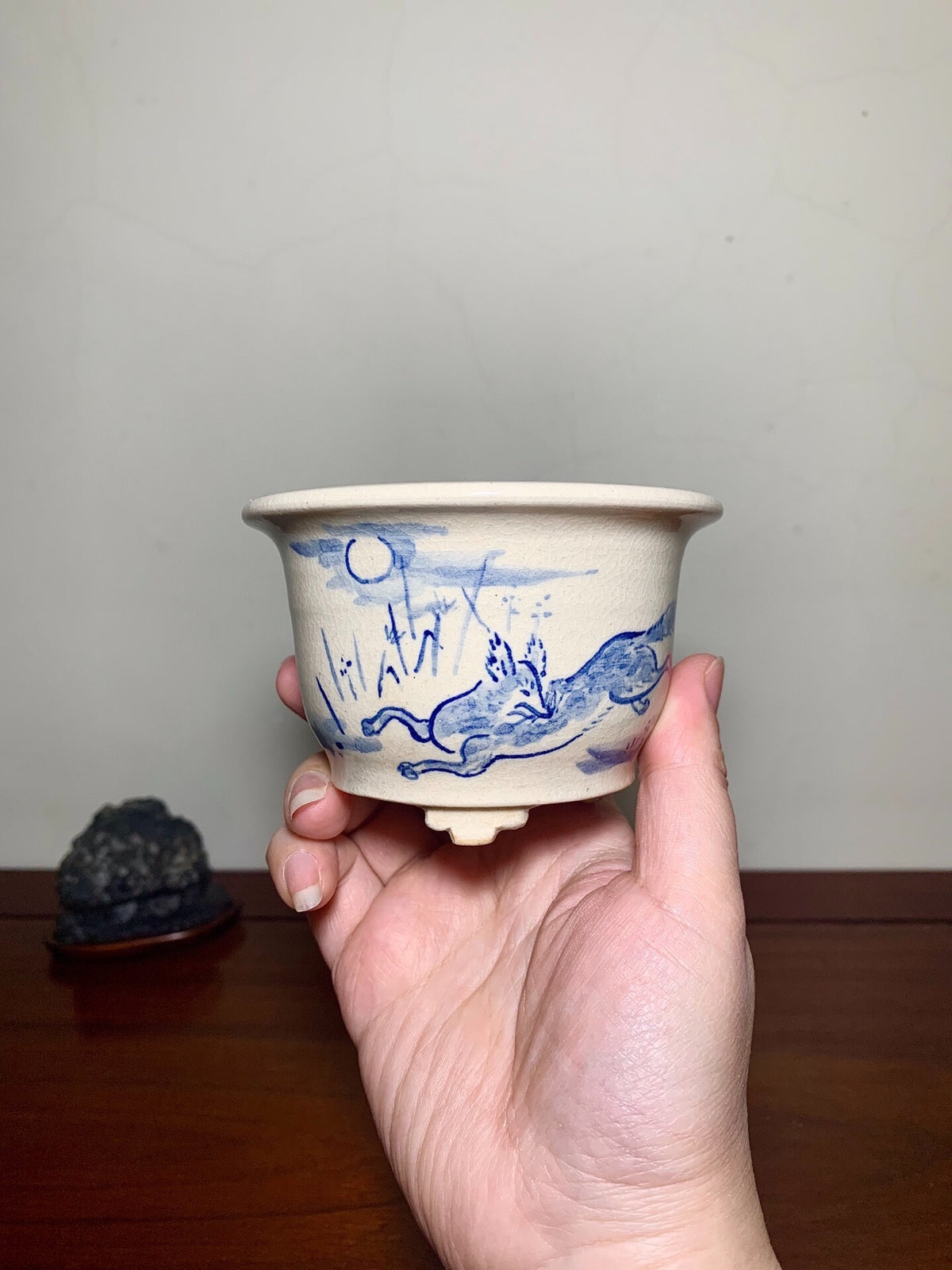 A hand holds a beige ceramic pot with blue painted landscape and rabbit design.