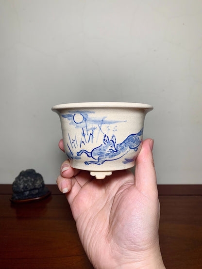 A hand holds a beige ceramic pot with blue painted landscape and rabbit design.