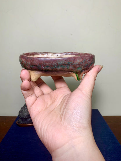 A hand holds a small, round, glazed ceramic pot with a marbled reddish-brown and green glaze.