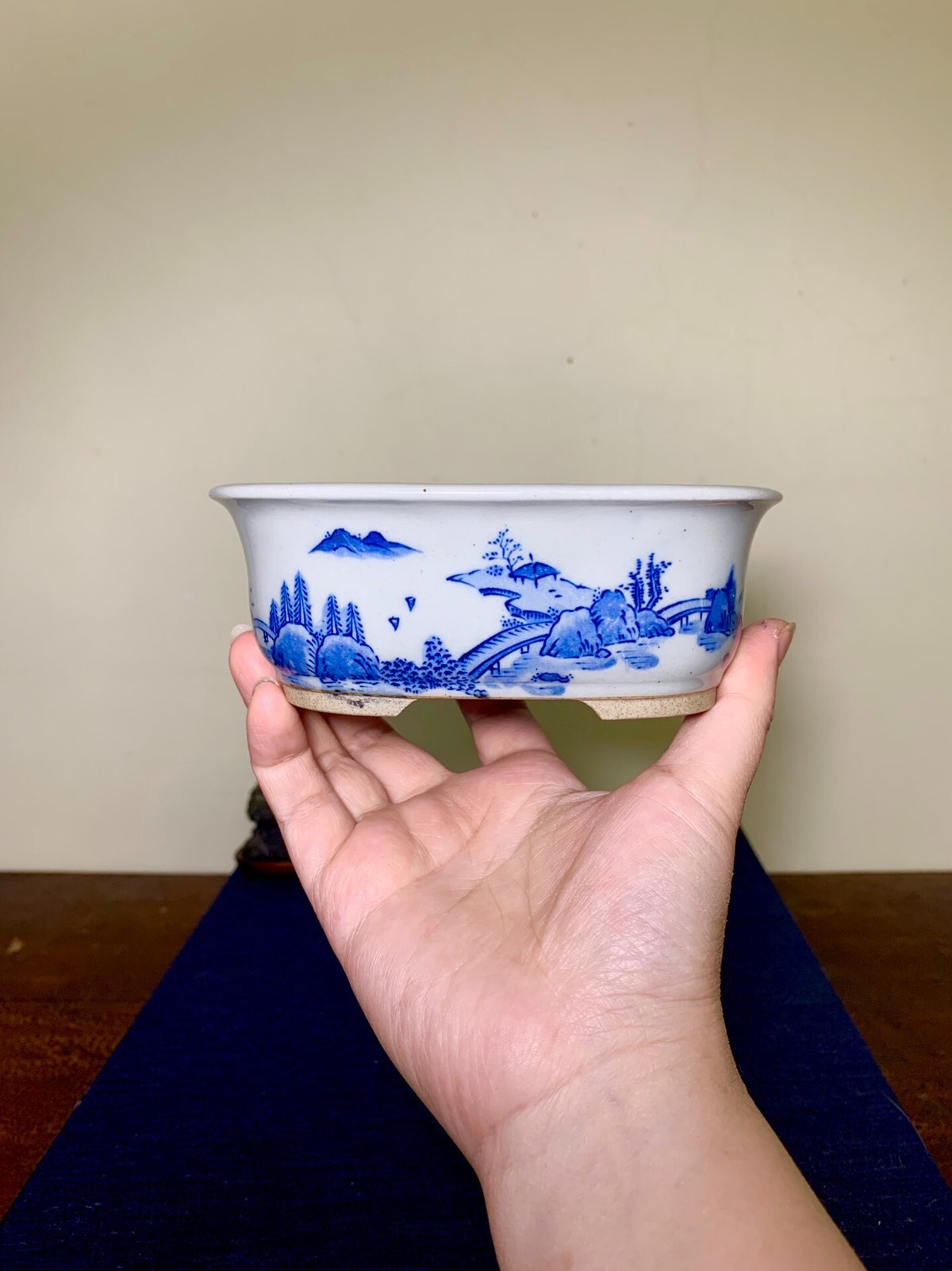 A hand holds a white glazed pot with blue landscape decoration.