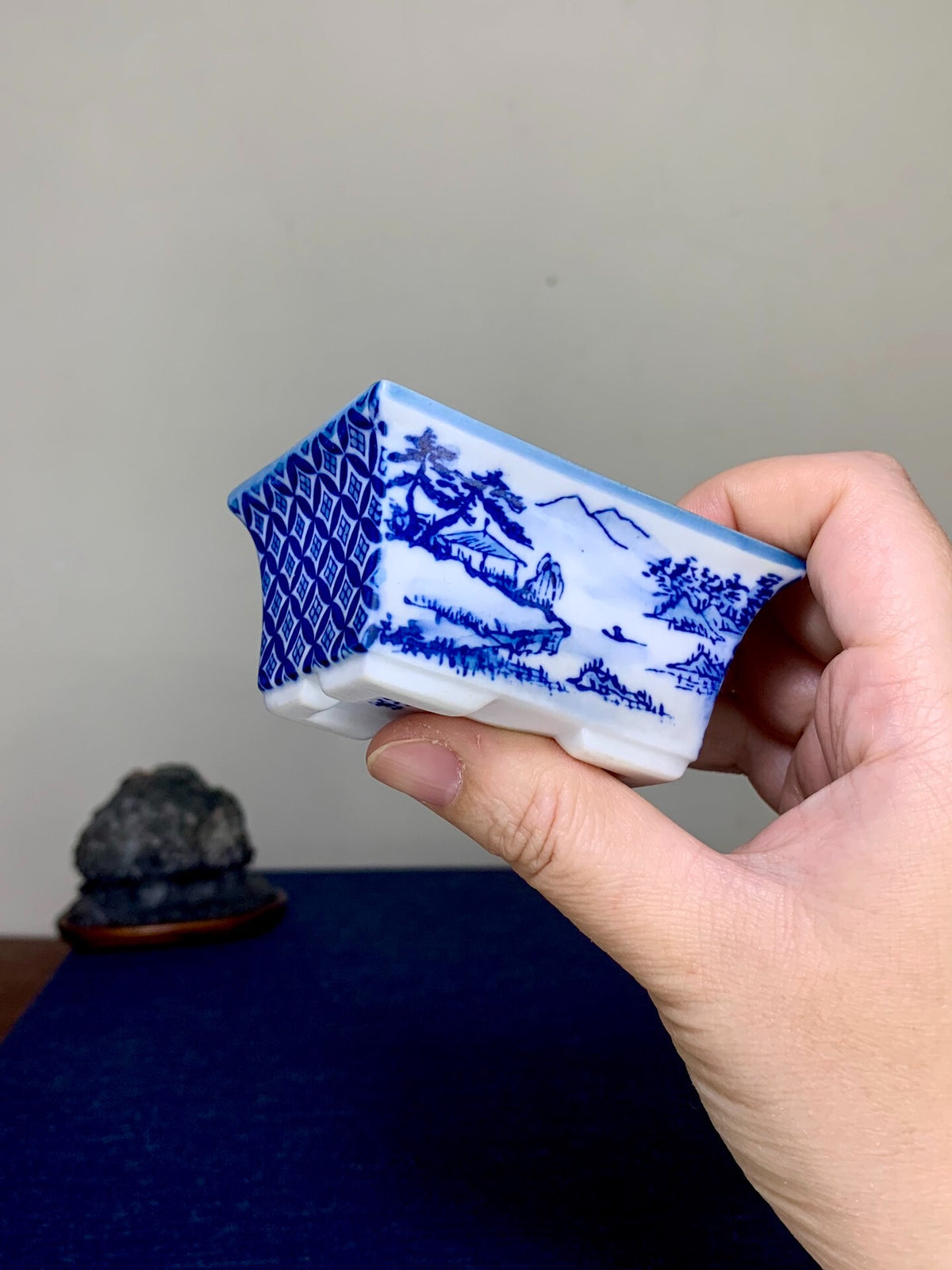 A hand holds a small, rectangular blue and white ceramic pot with landscape scenes and geometric patterns.