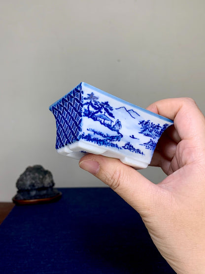 A hand holds a small, rectangular blue and white ceramic pot with landscape scenes and geometric patterns.