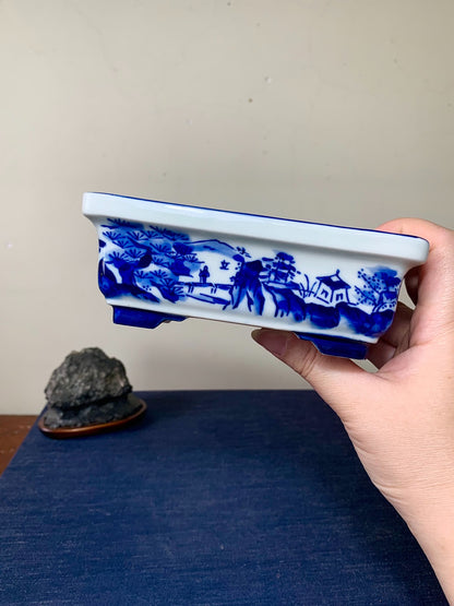 A hand holds a white rectangular pot with blue painted landscape scenes and a blue rim.