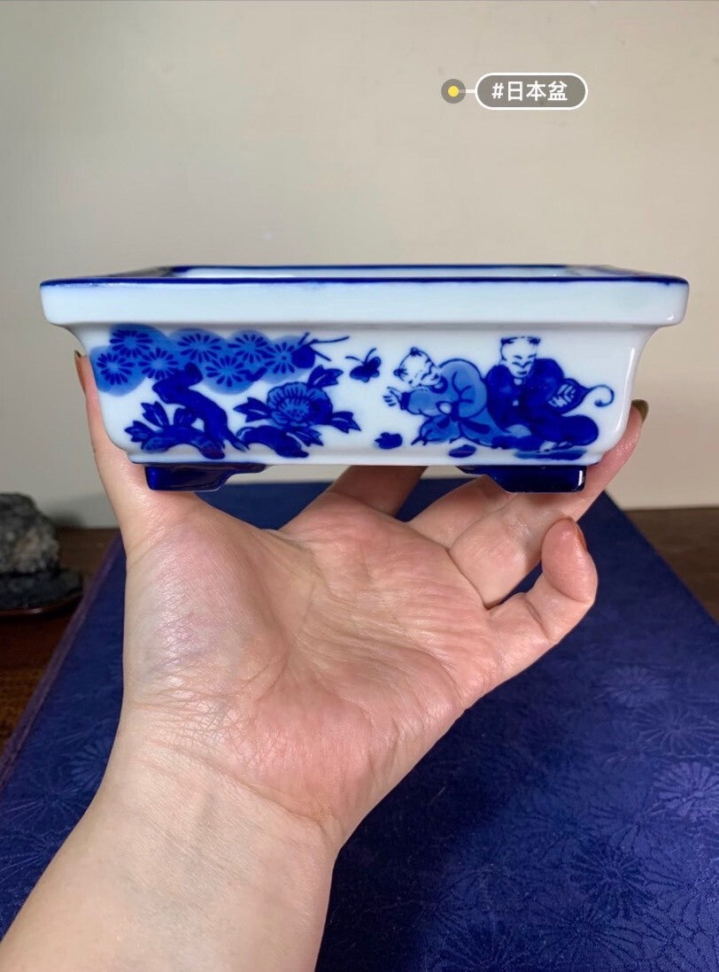 A hand holds a white rectangular pot with blue floral and child figures, glossy glaze.