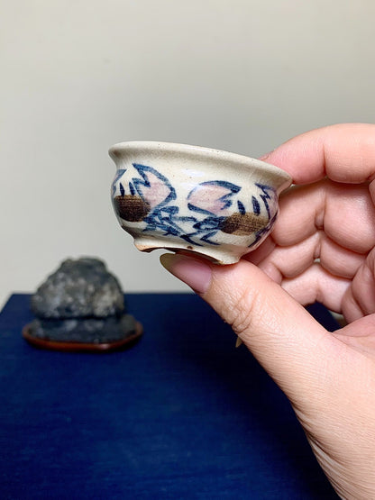 A hand holds a small, light-colored ceramic pot with blue and brown floral patterns.